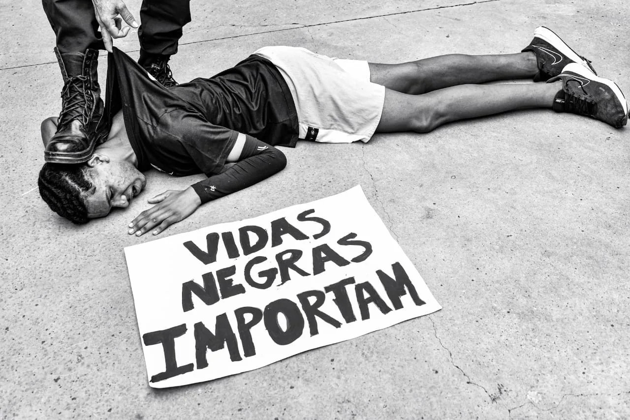 Cena de denúncia mostra um jovem no chão e um cartaz Vidas Negras Importam